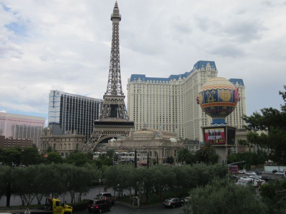 Las Vegas Strip scene showing Eiffel Tower replica, Paris Las Vegas hotel, and other buildings. Several cars are visible on the road