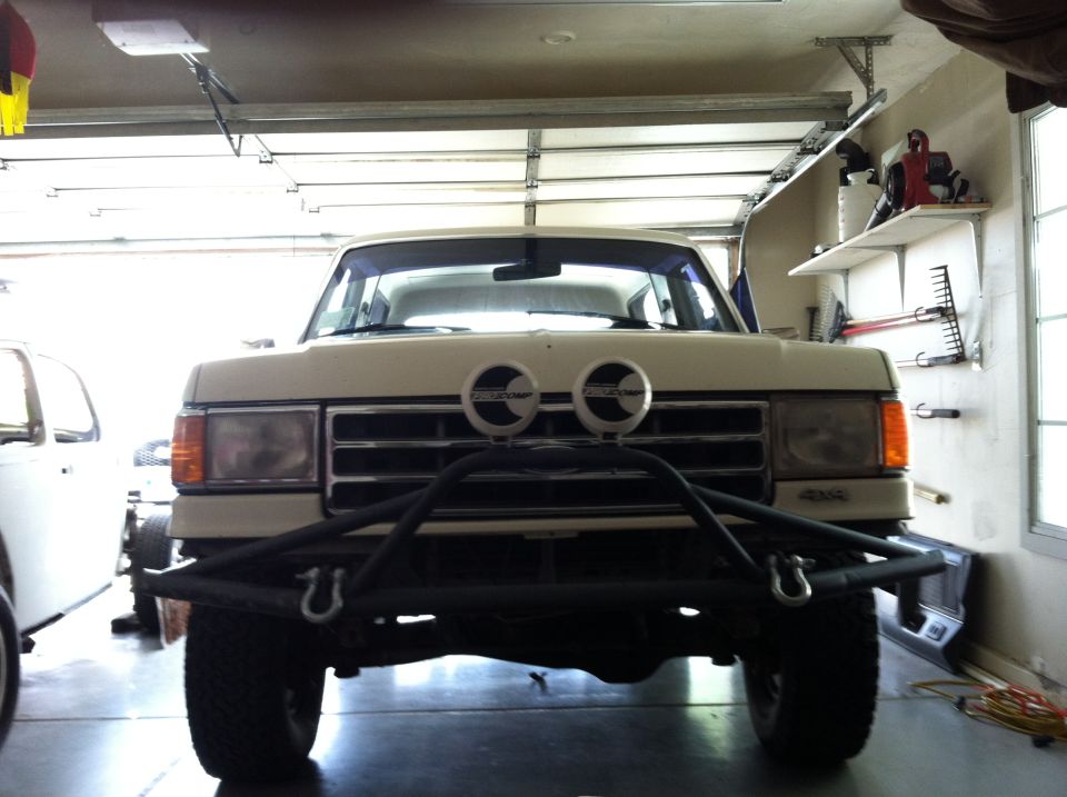 Front view of white lifted Ford Bronco in garage with black tube bumper and Pro Comp round lights