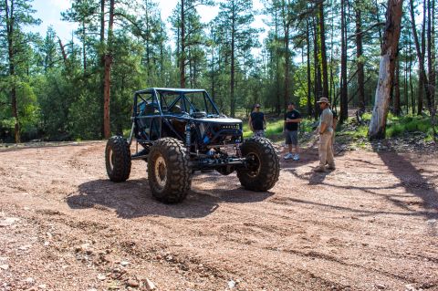 1966 Ford Bronco - A heavily modified off-road vehicle with a custom roll cage sits on a dirt path surrounded by trees.