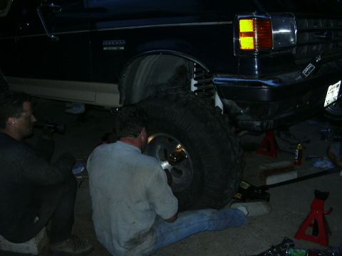 1991 Ford Bronco - Two men work on the lifted suspension and large tires of a black Ford Bronco.
