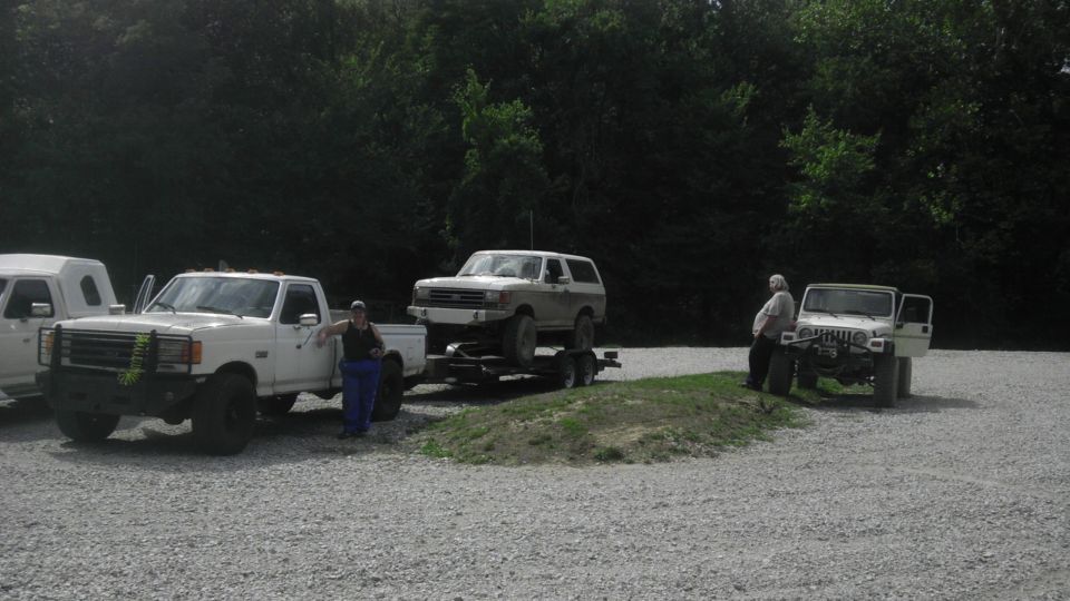 White Ford Bronco on a trailer pulled by a white pickup truck. Another white Jeep is parked nearby