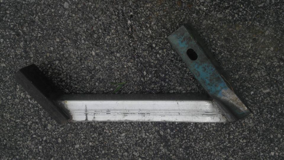 Two metal jack stands on asphalt. One is silver and L-shaped, the other is blue and wedge-shaped