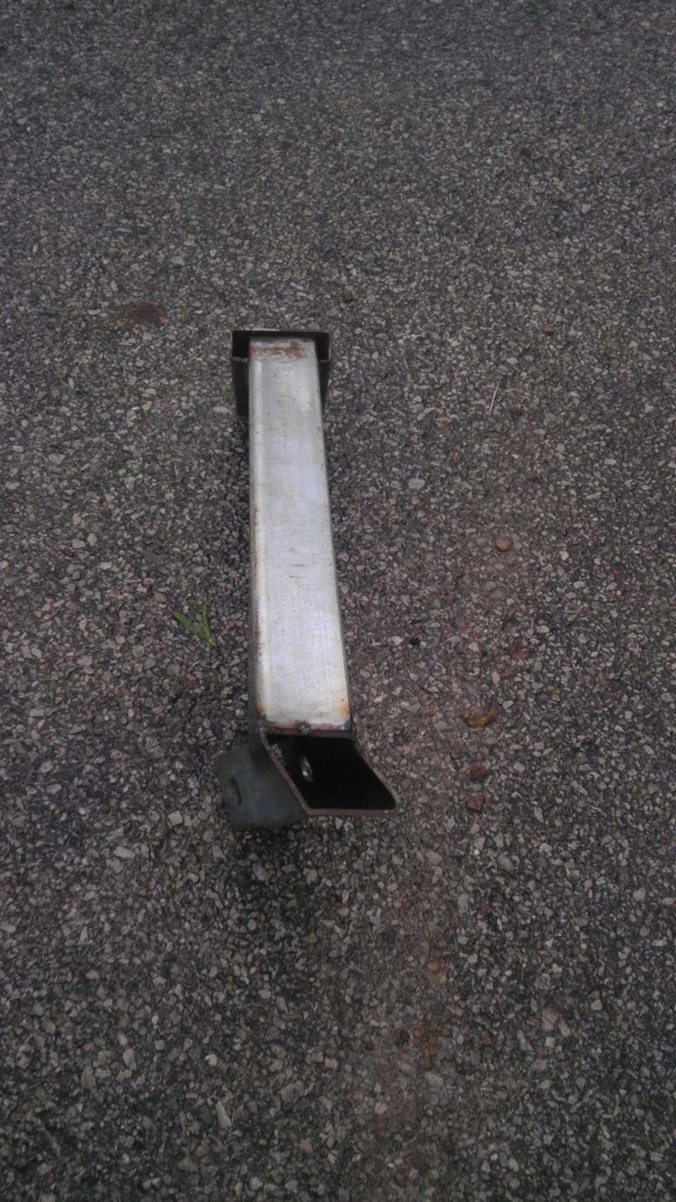 Metal bracket with a square tube and a flat base, lying on asphalt