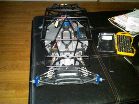 1986 Ford Bronco - RC car chassis assembly with roll cage, suspension, and electronics. Tools and papers are on the table.