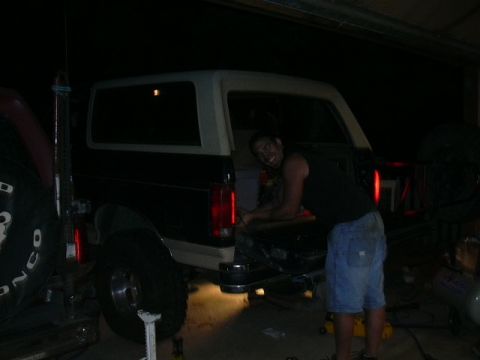 1991 Ford Bronco - Man in tank top and shorts working at the rear of a lifted black-over-white Ford Bronco inside a dimly lit garage.