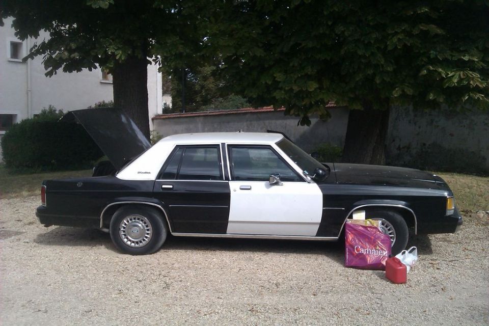 Black and white 1991 Ford Crown Victoria sedan parked outside with trunk open. Gas can and shopping bag on the ground