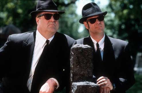 Two men in black suits, white shirts, black ties, and fedoras stand outdoors. Both wear sunglasses