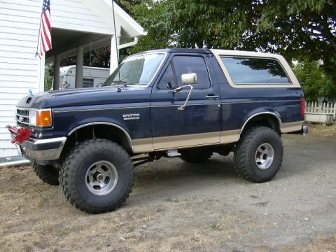 1991 Ford Bronco - Dark blue two-tone Ford Bronco with tan lower body cladding and matching tan hardtop, shot from the front three-quarter angle in a gravel d…