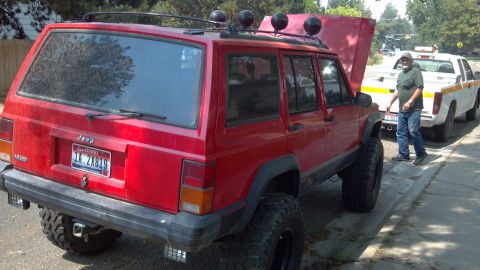 1995 Jeep Cherokee - A red 1995 Jeep Cherokee is seen from the rear, aftermarket lights and a roof rack, with another vehicle and a person in the background.