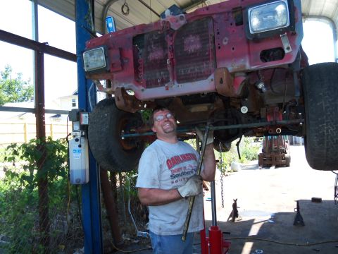 1987 Ford Ranger - Mechanic works on the front end of a red truck chassis elevated on a lift in an outdoor garage setting.