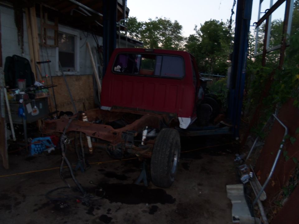 Red pickup truck body on a rusty chassis, raised on jack stands in a workshop