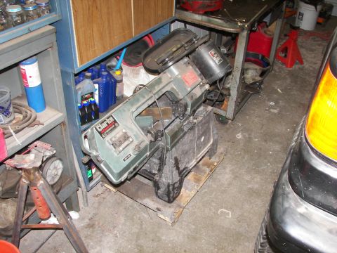 1988 Ford Bronco - A bandsaw sits on a pallet in a cluttered garage, with shelves of supplies and part of a vehicle in the foreground.