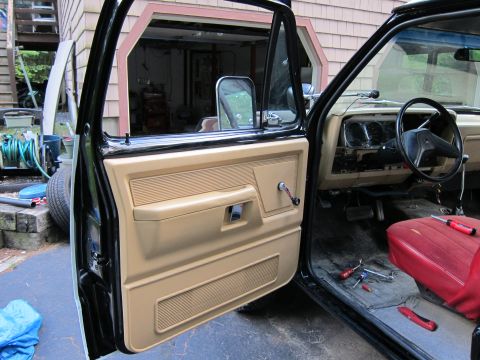 1987 Dodge 1500 - The interior of a truck is shown with the driver's door open, revealing the tan door panel and red seat.