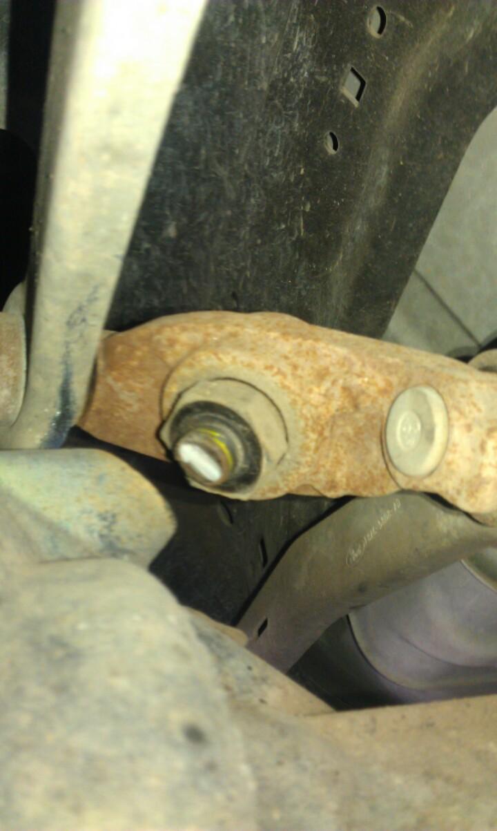 Underside view of a rusty suspension component with a bushing bolt, photographed beneath a vehicle