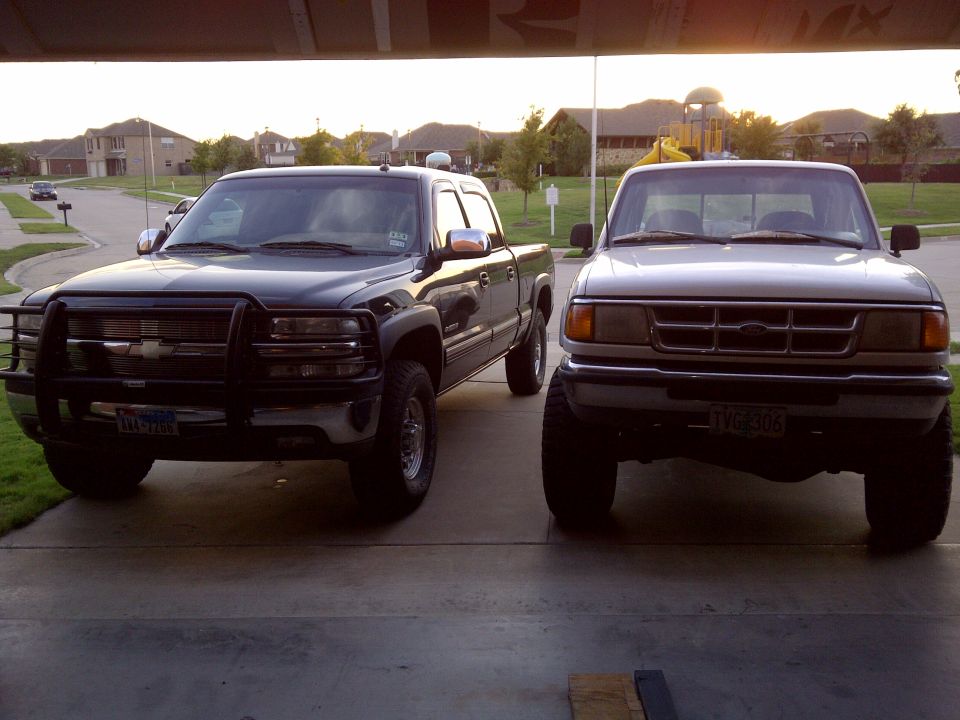 Two pickup trucks parked side-by-side. The truck on the right is a silver Ford Ranger. The truck on the left is a dark-colored Chevrolet