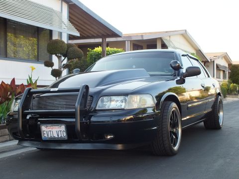 2003 Ford Crown Victoria - Front three-quarter view of a blacked-out Crown Victoria sedan wearing a push bumper and black multi-spoke wheels.