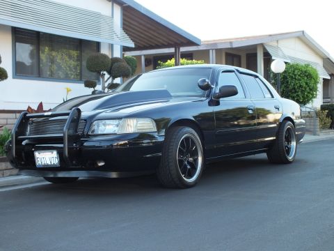 2003 Ford Crown Victoria - Side view of the blacked-out sedan parked at a curb showing its long body and dark tint.