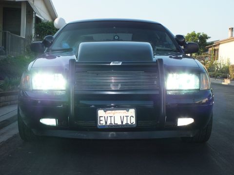 2003 Ford Crown Victoria - Head-on dusk shot of the sedan with all lights on, showing grille guard and EVIL VIC plate.