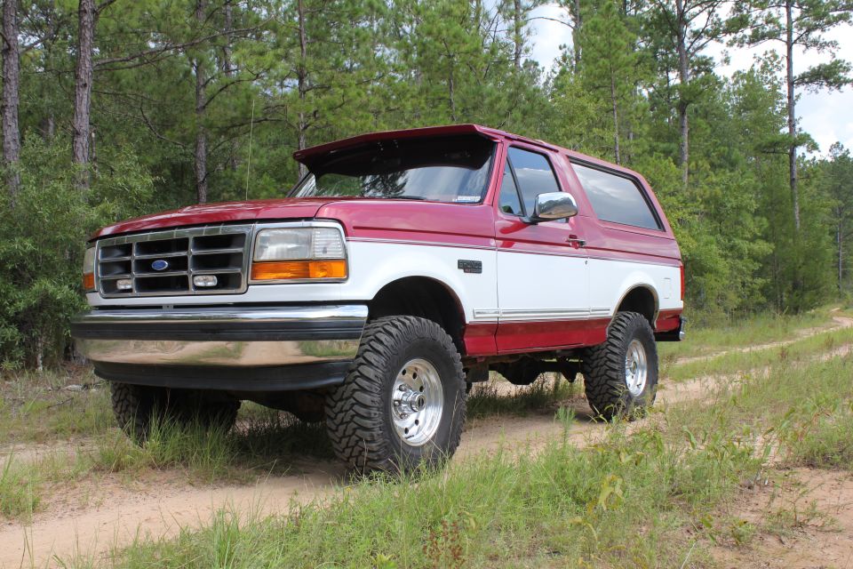 Red and white Ford Bronco on a dirt road with trees in the background