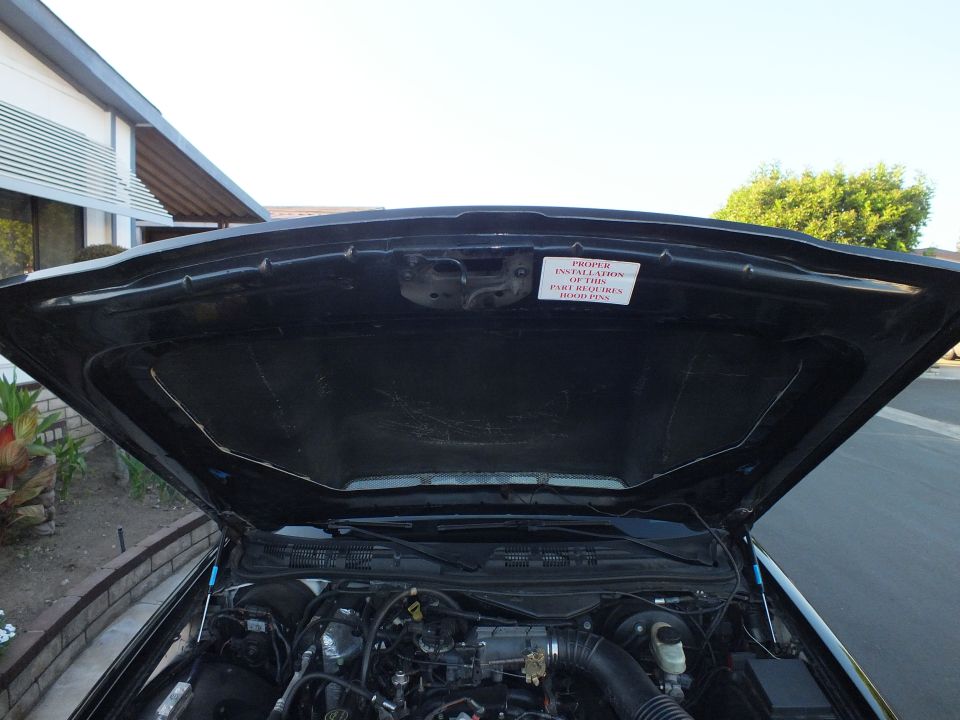 Open hood underside of black sedan with hood-pin warning sticker over engine bay