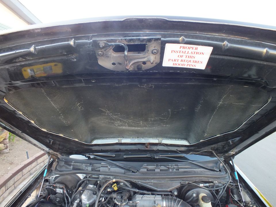 Underside of black sedan hood with scratched liner and hood-pin install warning label