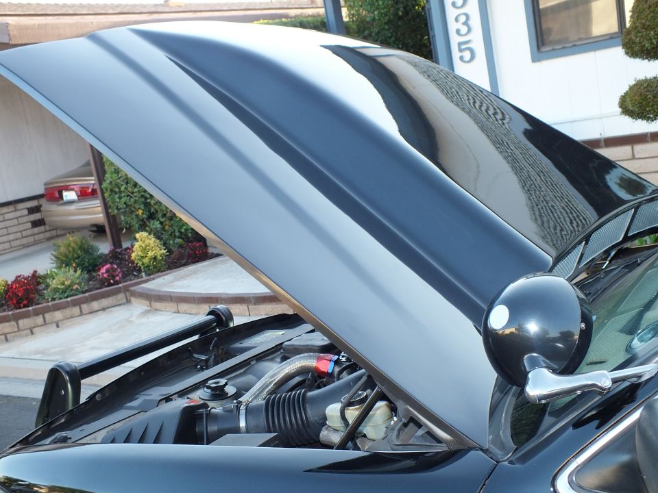Open black hood of dark sedan over engine bay with circular side mirror in driveway