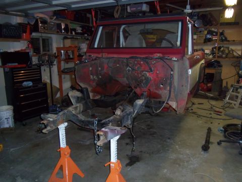1968 Ford Bronco - The body of a red SUV is elevated on jack stands in a garage, with its engine and front end disassembled.