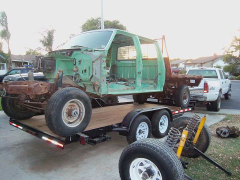 1968 Ford Bronco - A green truck cab and chassis is loaded onto a trailer, towed by a white pickup truck, with spare parts nearby.