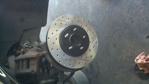 2003 Ford Crown Victoria - Cross-drilled slotted brake rotor mounted to a five-stud hub, caliper visible behind on a raised car.