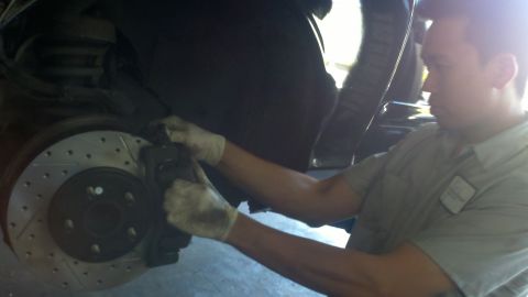 2003 Ford Crown Victoria - Gloved technician in tan shirt services a slotted brake rotor and caliper beneath a lifted car.