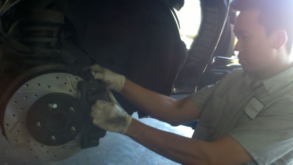 Mechanic in gloves working on drilled brake rotor and caliper under raised vehicle