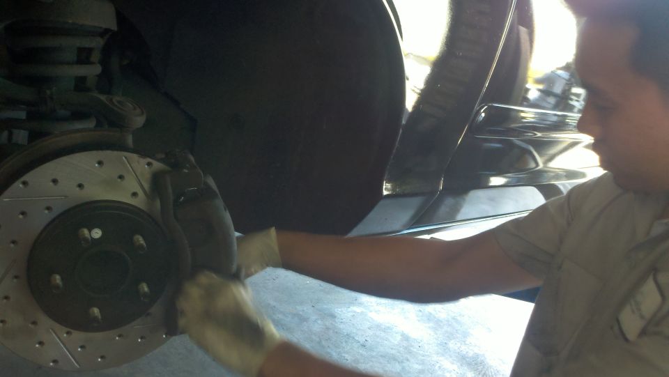 Mechanic reaching into wheel well with drilled rotor and coil spring on lifted car