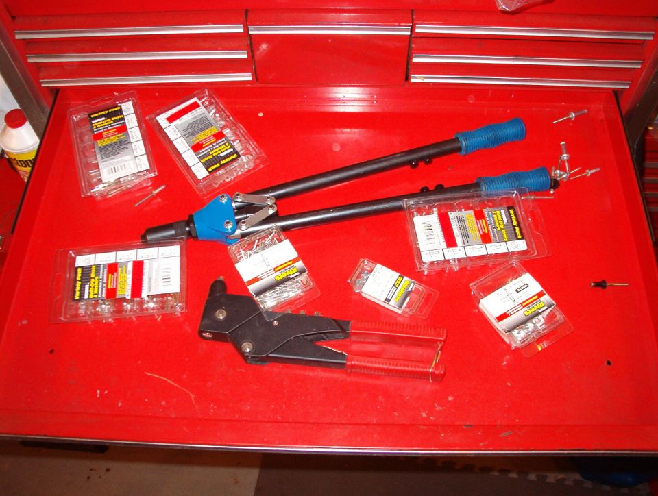 Rivet gun with blue handles and packages of assorted rivets on a red toolbox surface