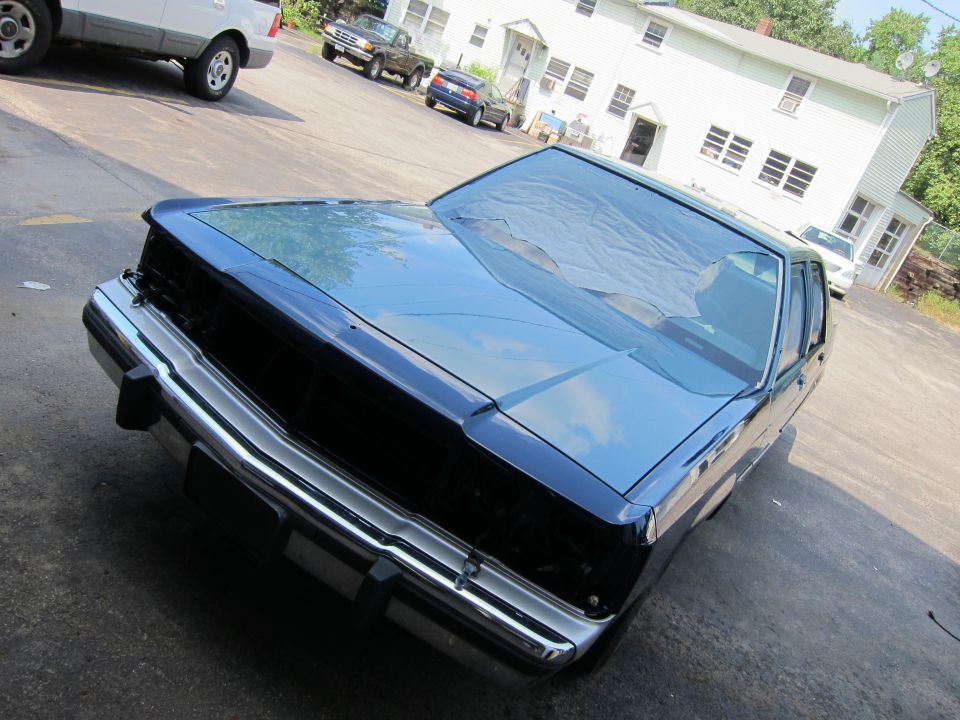 Dark sedan with chrome bumper and grille, parked outside a white house. Another SUV and pickup truck are visible