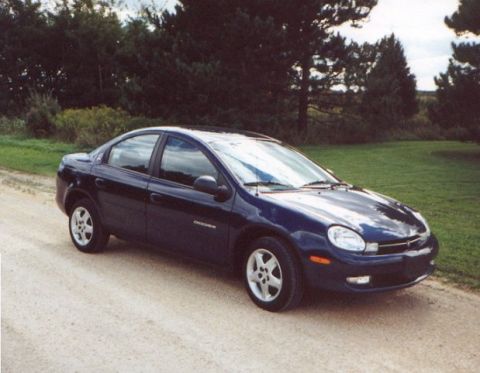 2000 Dodge Neon - A dark blue 2000 Dodge Neon sedan is parked on a dirt road with trees in the background.