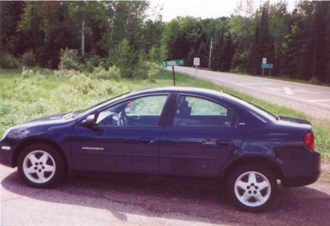 2000 Dodge Neon - A dark blue 2000 Dodge Neon sedan is parked on the side of a rural road with trees and a "S HOOL" sign in the background.