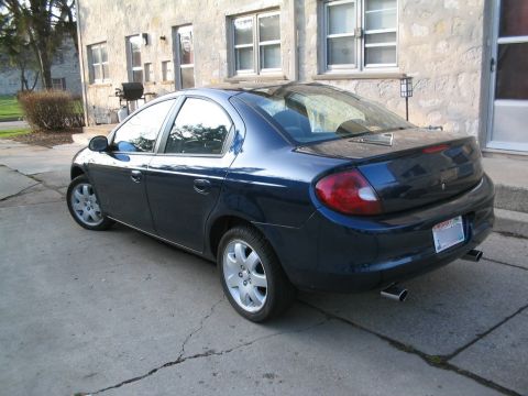 2000 Dodge Neon - A dark blue 2000 Dodge Neon sedan is parked on a cracked concrete driveway in front of a stone building.