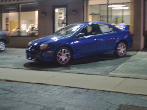 2000 Dodge Neon - A blue sedan with a spoiler is parked in front of a building. The vehicle appears to be a 2000 Dodge Neon.