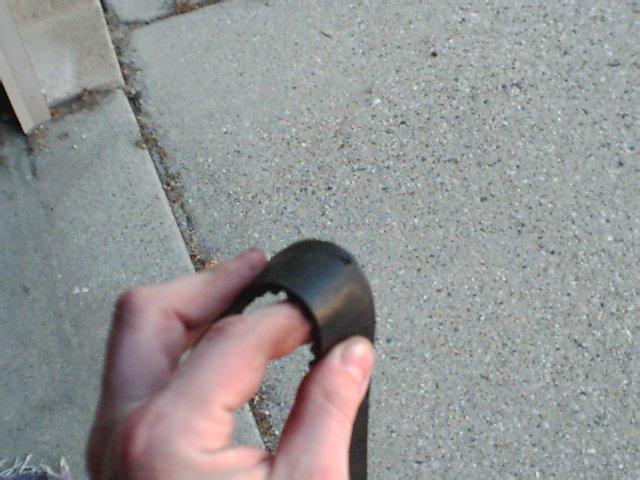 Hand holding a black plastic automotive part against a concrete and paver background. Text 'SUPERMOTORS' is visible