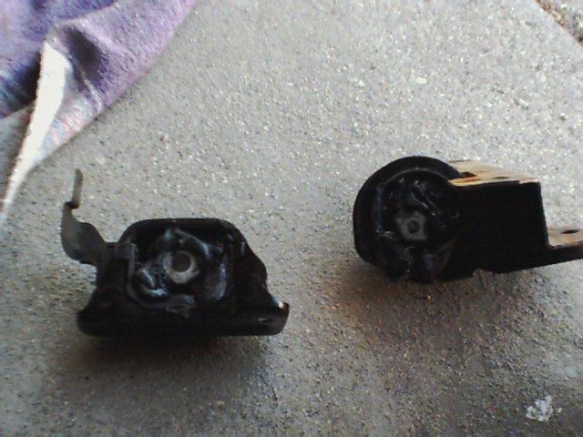 Two black engine mounts on a concrete surface, one with a metal bracket attached