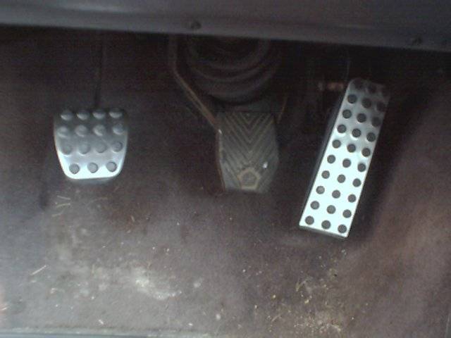 Car interior showing three pedals: clutch, brake, and accelerator. The accelerator pedal has a perforated metal cover
