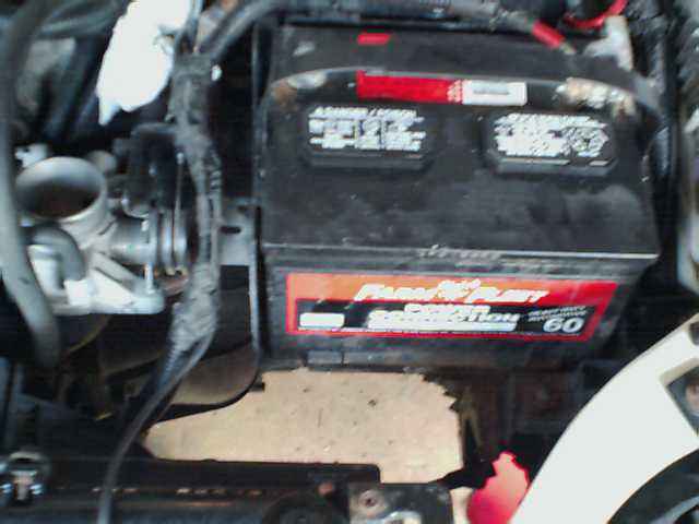 Car battery with red and black casing, labeled "Flow-Rite", and engine parts including a metal tube and wiring