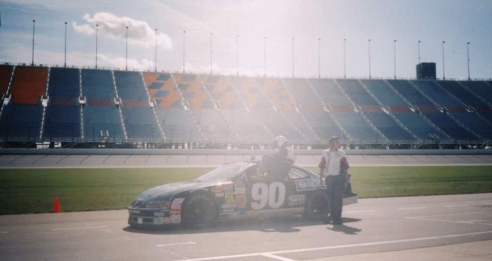 Stock car #90 parked on a racetrack in front of stadium seating