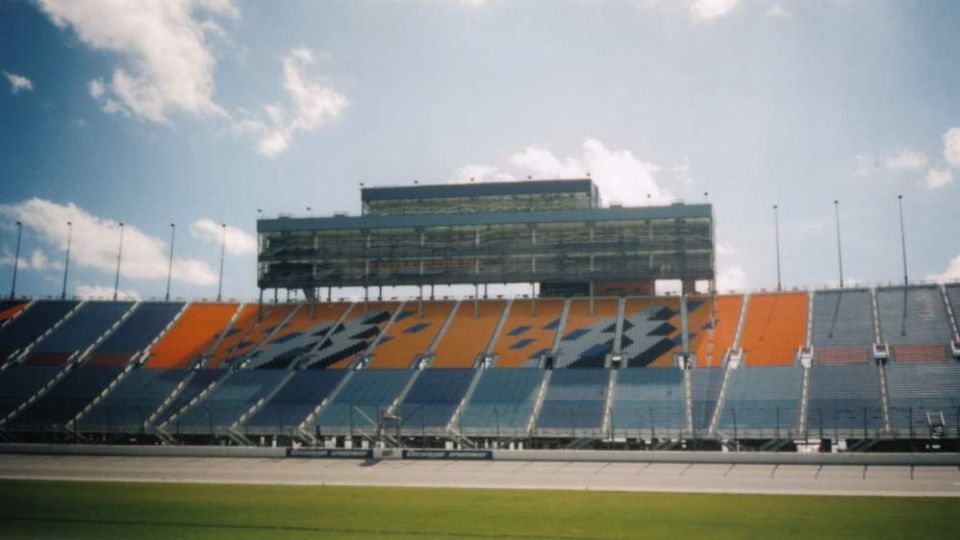Racetrack grandstands with blue and orange seats under a cloudy sky. A large building with multiple levels is visible at the top