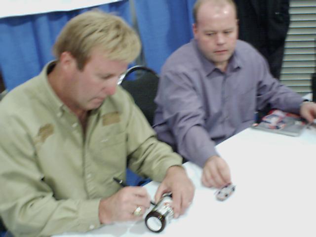 Two men sit at a white table, one signing a bottle, the other holding a toy car