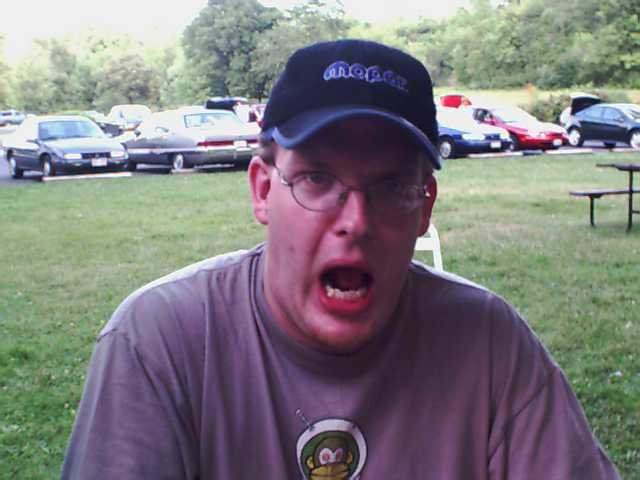 Man in a cap and t-shirt with mouth open, cars parked in background