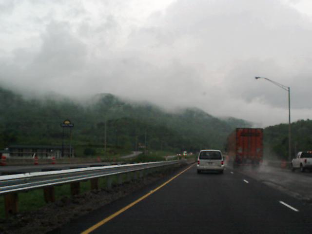 Highway with a white van, red truck, and white pickup truck under cloudy skies and misty mountains