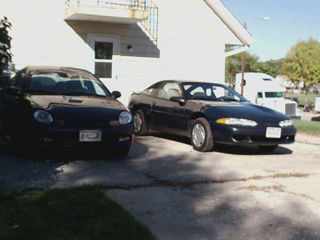 Two dark sedans parked in front of a white building. A semi-truck is visible in the background