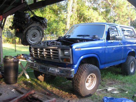 1994 Ford Bronco - A blue Ford Bronco is parked outside with its hood open as an engine is hoisted above it.