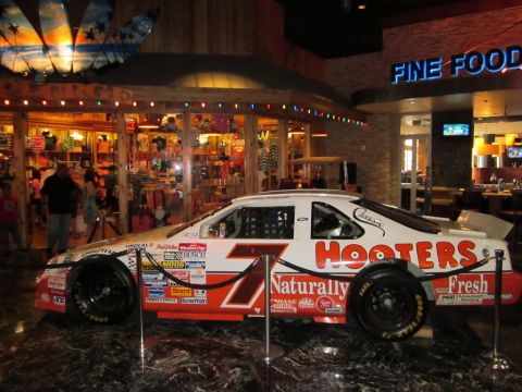 1991 Ford LTD Crown Victoria - Hooters #7 race car on display indoors, with "FINE FOOD" sign visible in the background.